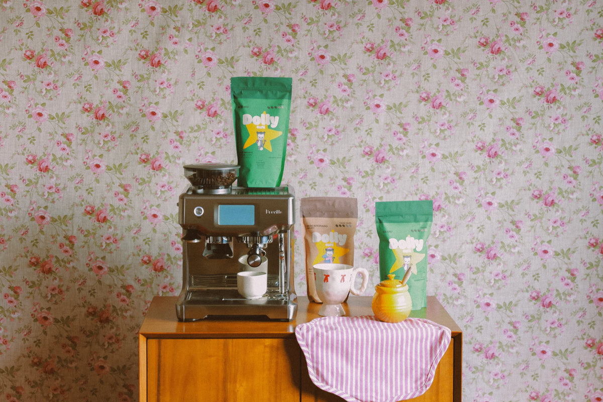 Café Dolly junto a una máquina de espresso sobre un mueble de madera, con taza y papel tapiz floral